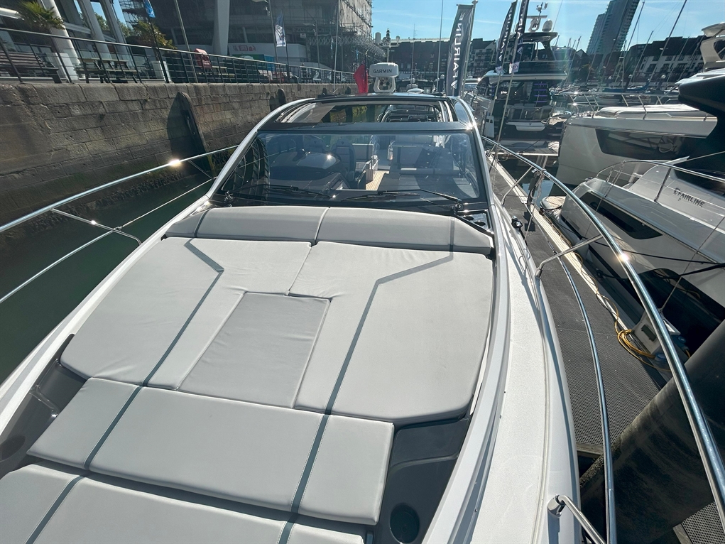 A sleek motorboat's deck with sunbathing cushions, showing a clear sky and a marina with other boats in the background.